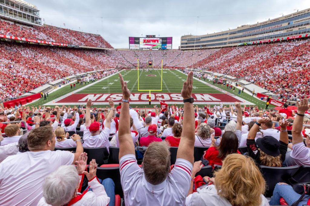 A packed college football stadium with fans in red and white attire cheering and raising their arms in excitement, displaying a vibrant atmosphere during a game.